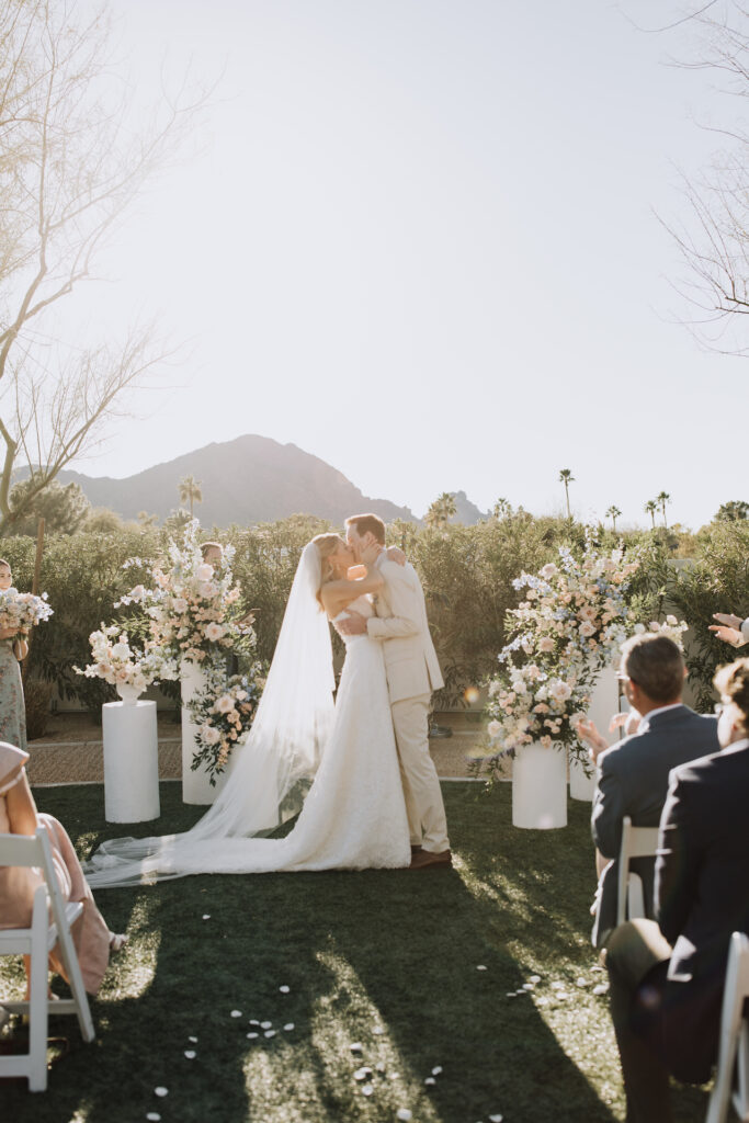 Bride and groom kissing in outdoor wedding ceremony altar space at Andaz Resort with flowers placed on pillars on either side of them and Camelback Mountain in backgrounnd.