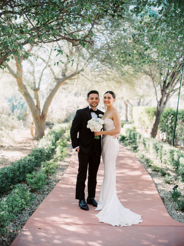 Bride and groom standing close on path at El Chorro, both smiling, bride holding bouquet of white flowers.