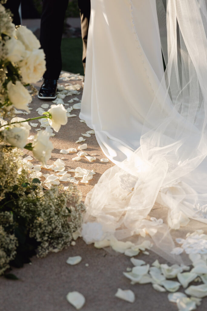 Bride's veil trailing behind her on ground with white rose petals sprinkled about.