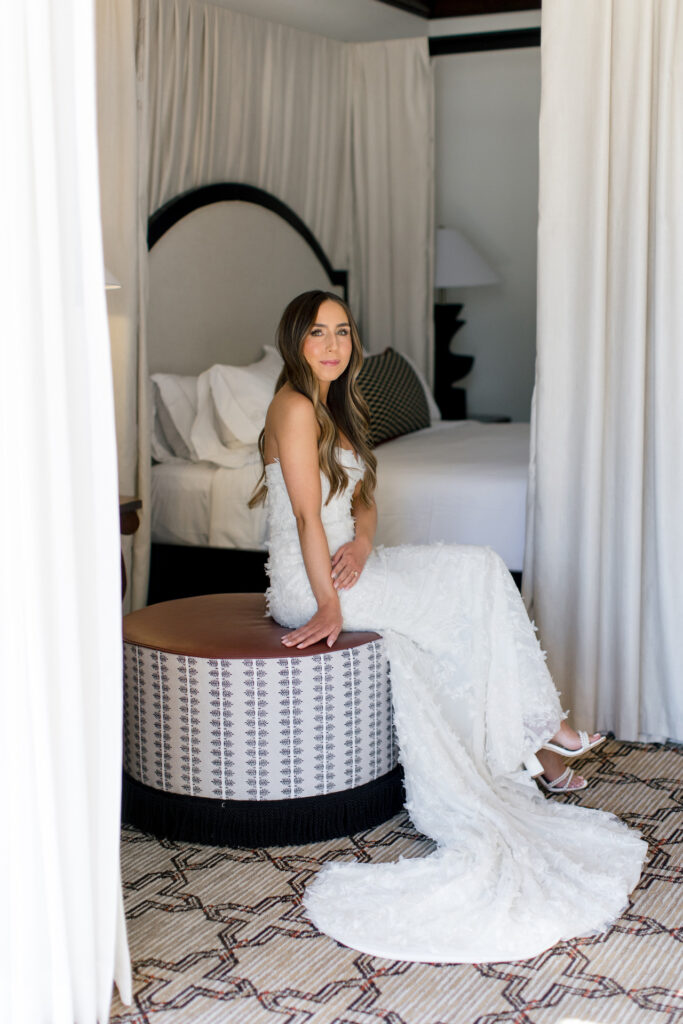 Bride sitting in round ottoman next to bed, smiling, in resort room.