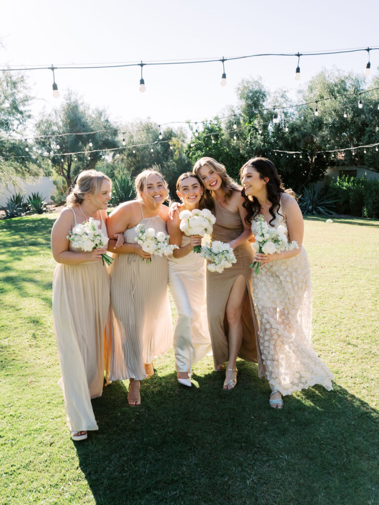 Bride walking with bridesmaids, all smiling. Bridesmaids wearing different design dresses in shades of cream and tan.