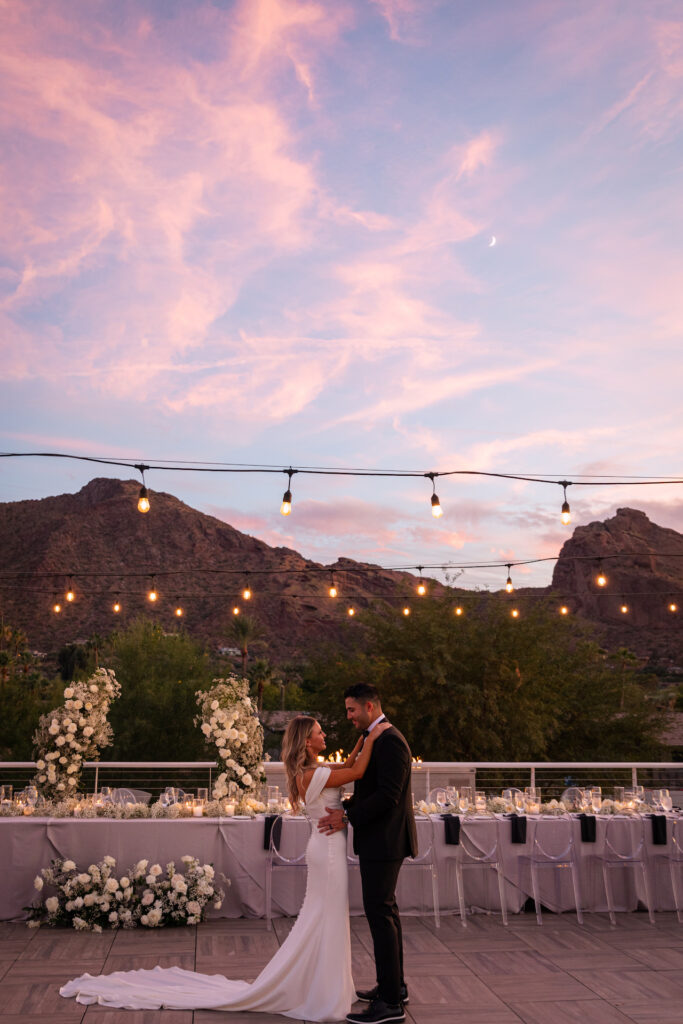 Bride resting her hands and groom's shoulder's, facing each other on Mountain Shadows patio set for outdoor wedding reception with Camelback Mountain in background.