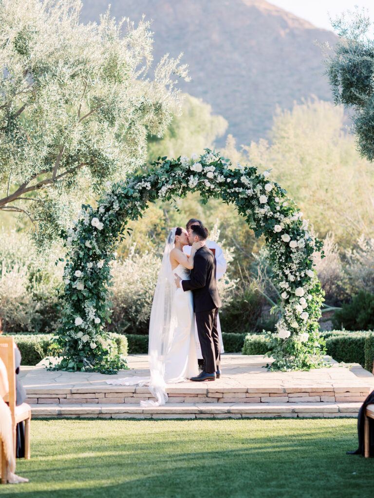 Bride and groom kissing under wedding ceremony arch of greenery and white flowers.