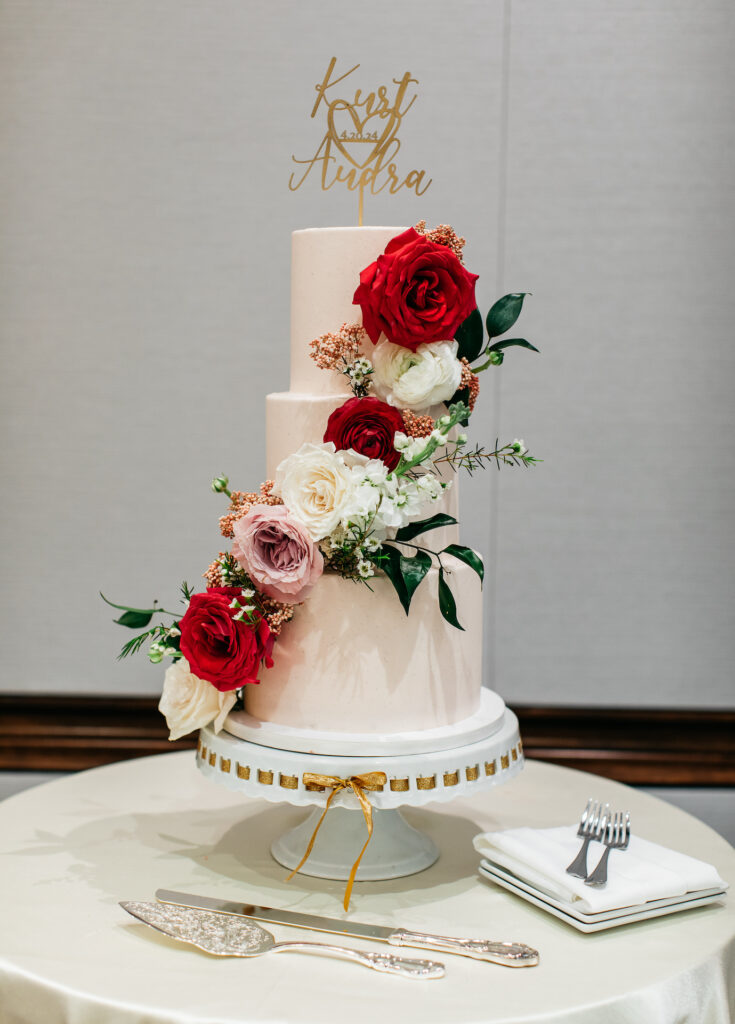 White tiered wedding cake with white, red, and pink roses placed up its side.