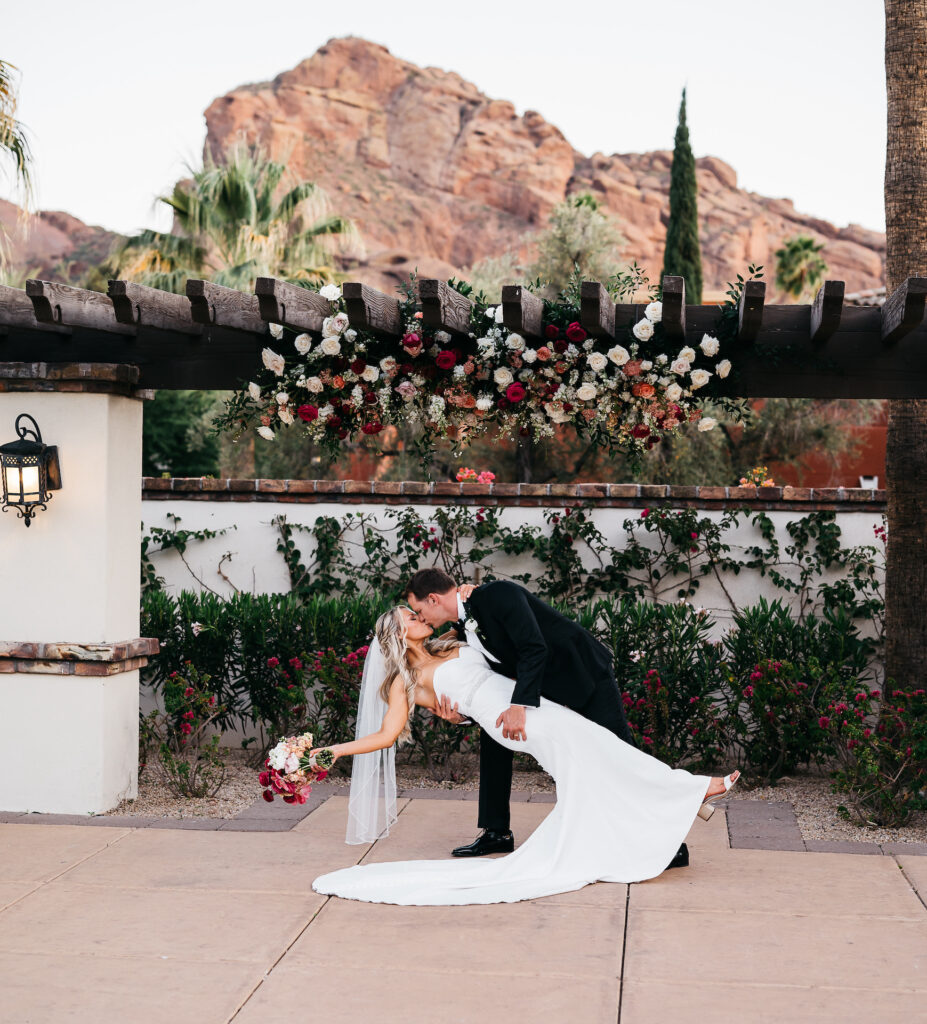 Groom dipping bride and kissing her while she holds her bouquet off to side under pergola with flowers installed on it.