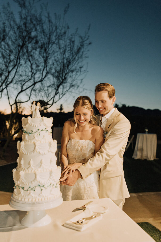 Bride and groom cutting three tiered white wedding cake together with piping details.