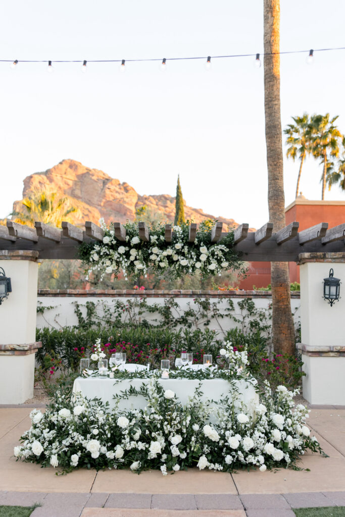 Reception head table with white flowers and greenery design above on pergola, on table, and on ground in front.