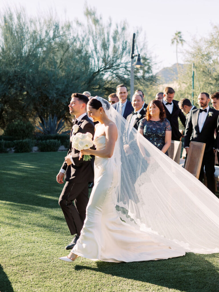 Bride walking with man in black suit down ceremony aisle holding her bouquet of white flowers.