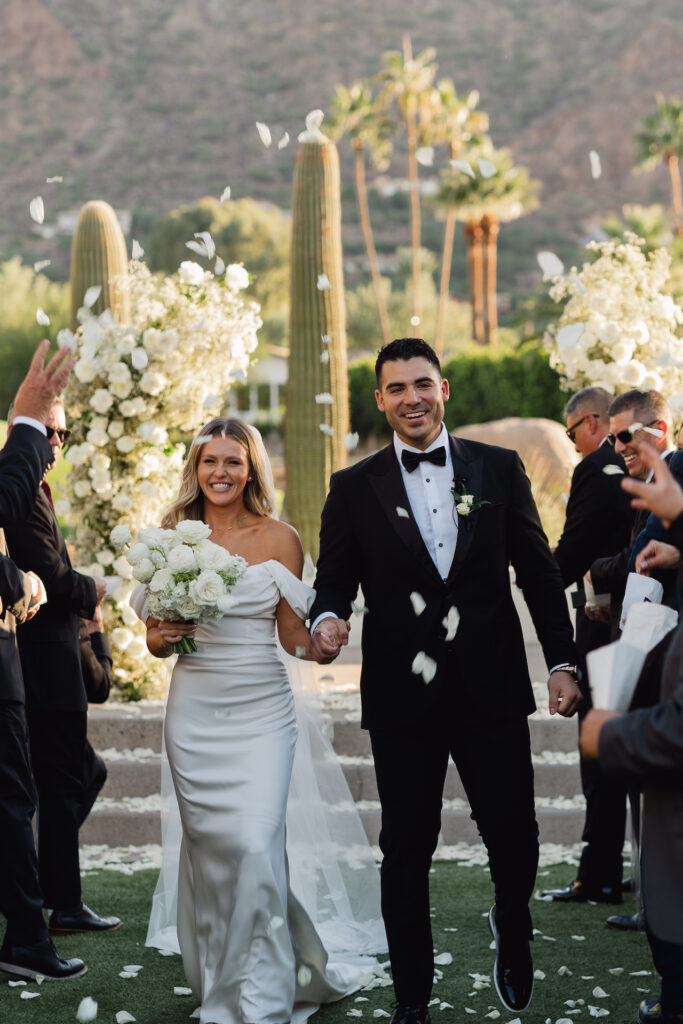 Bride and groom smiling, holding hands, walking down wedding ceremony aisle with white rose petals being tossed around them.