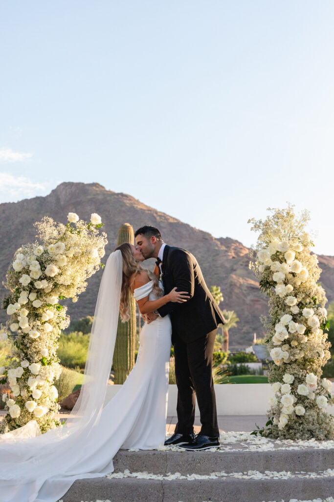 Bride and groom kissing in ceremony altar space at Mountain Shadows between two white floral columns.