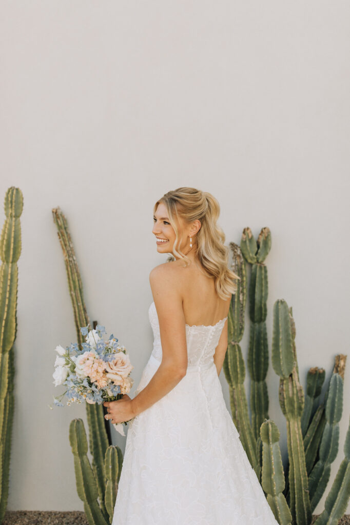 Bride turned away, looking over her shoulder and smiling while holding bouquet in hand standing in front of white wall at Andaz with cacti growing in front of it.