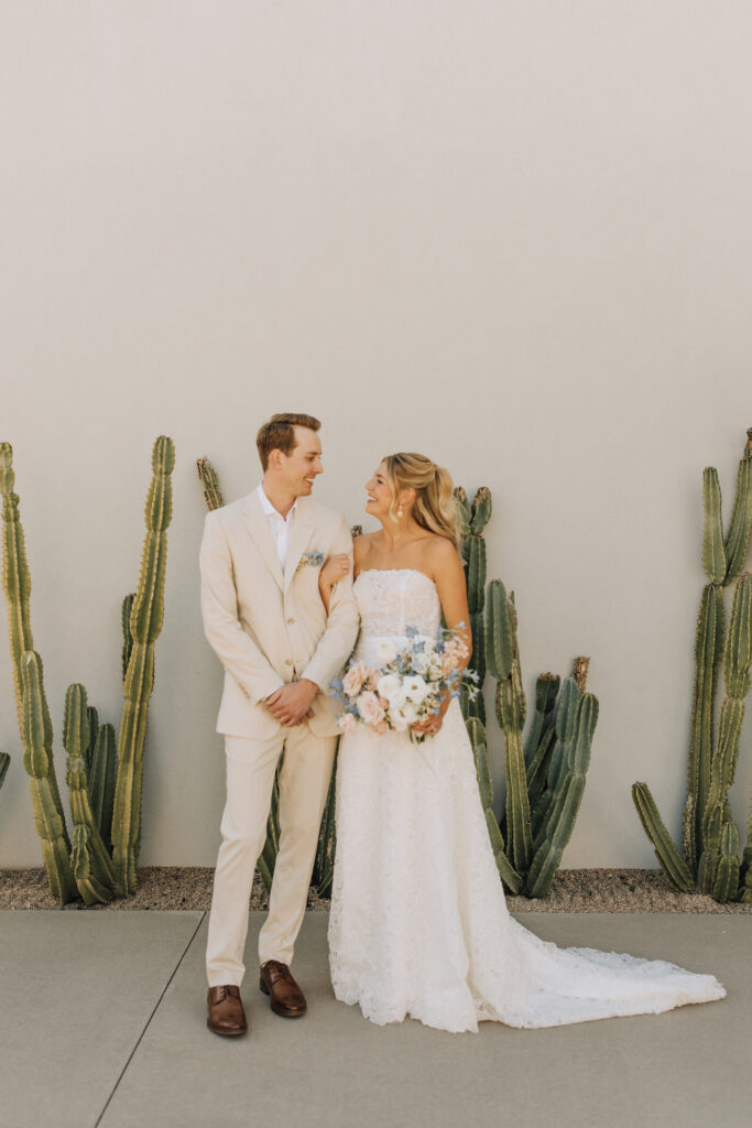 Bride with arm around groom's both smiling at each other, standing in front of white wall at Andaz with cacti growing in front of it.