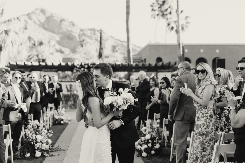 Bride and groom kissing at back of wedding ceremony aisle with guests clapping around them.