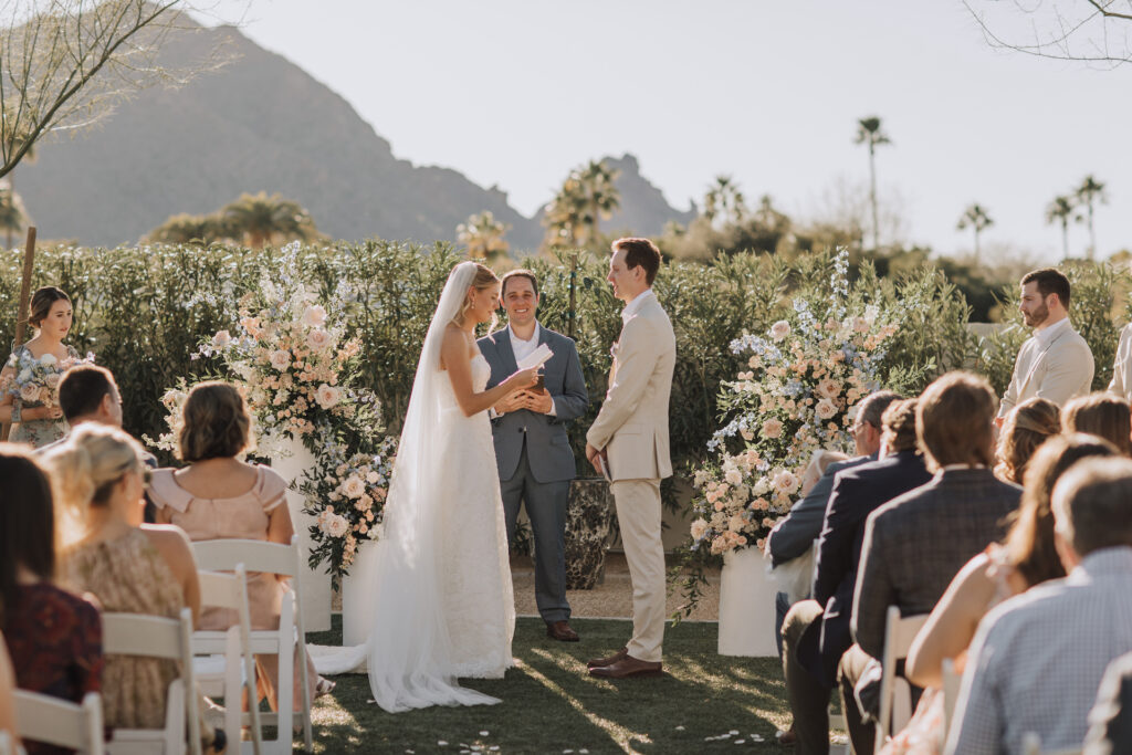 Bride and groom during wedding ceremony, bride reading vows to groom with guests watching on and officiant standing behind them.