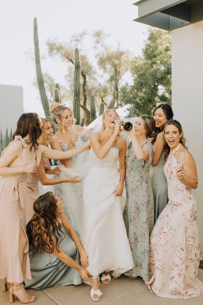 Bride with bridesmaids acting silly with champagne bottle to bride's mouth, bridesmaids adjusting her shoes and veil.