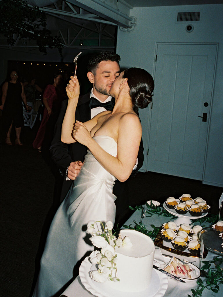 Bride and groom kissing at dessert table next to wedding cake.