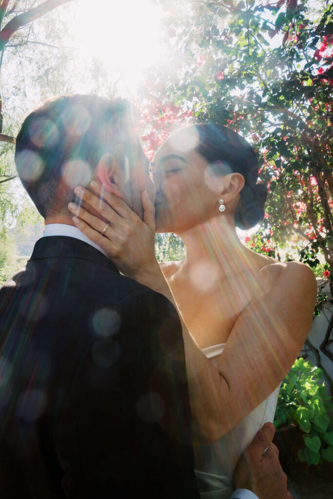 Bride and groom kissing under trees with sun rays coming through the leaves.