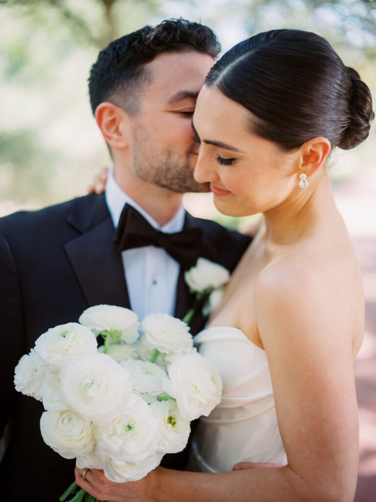 Groom kissing bride on cheek, bride smiling with eyes closed and holding bouquet of white ranunculus flowers.