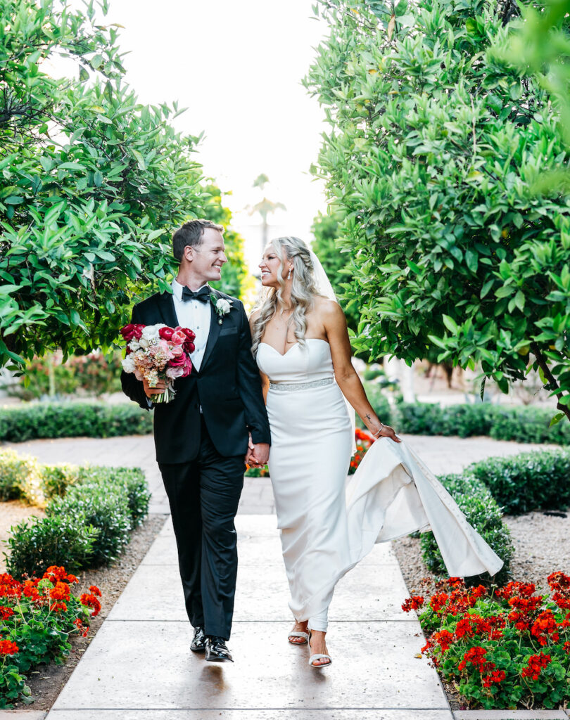 Bride and groom walking on path at Omni Montelucia resort holding hands, groom holding bouquet, both smiling.