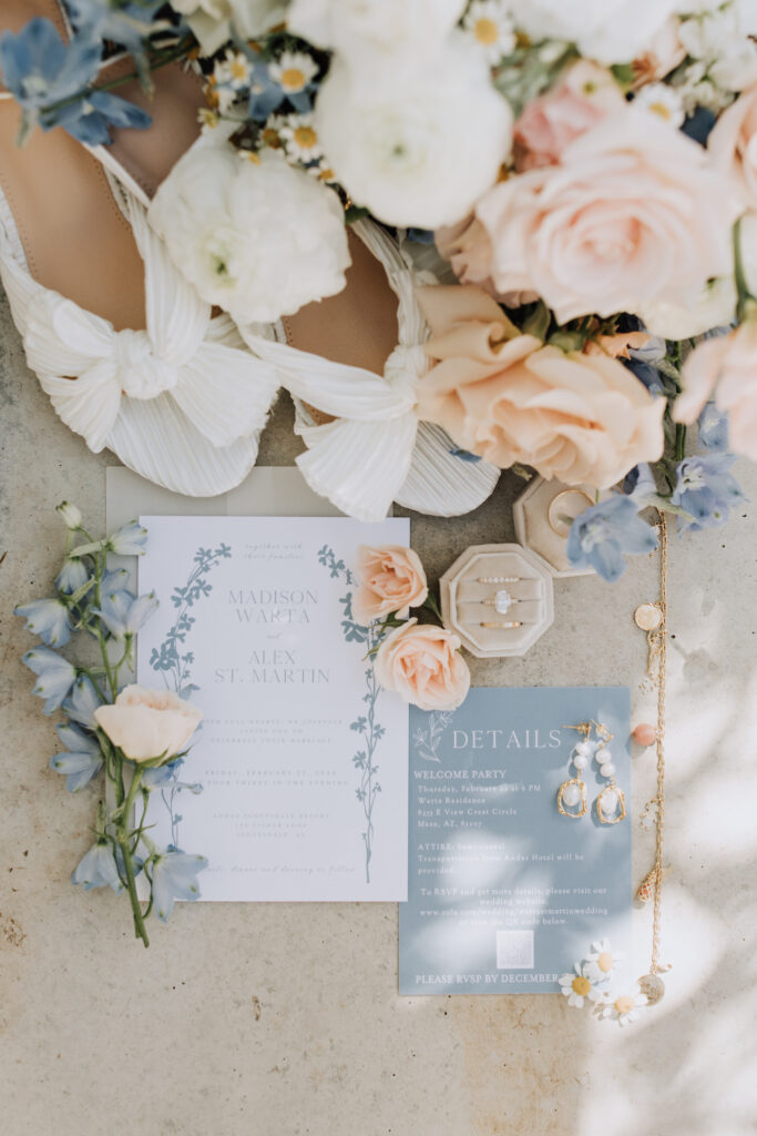 Wedding invitation flat lay of blue and white invitation, wedding rings, white bridal shoes, and bridal boquet.