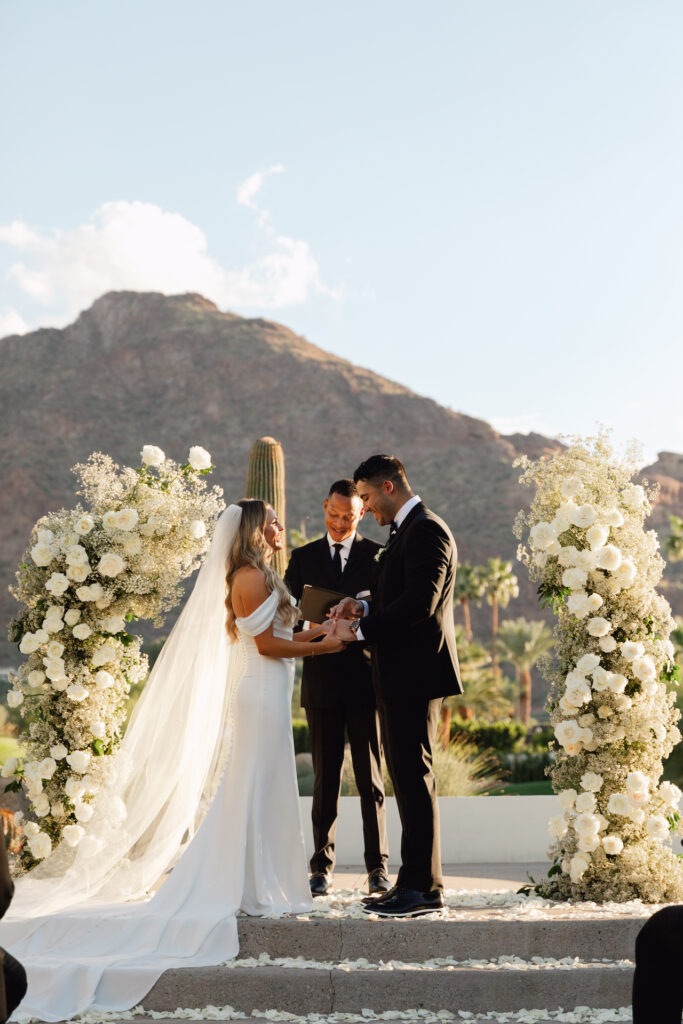 Bride and groom holding hands, smiling during outdoor wedding ceremony with officiant behind them and Camelback Mountain in background.