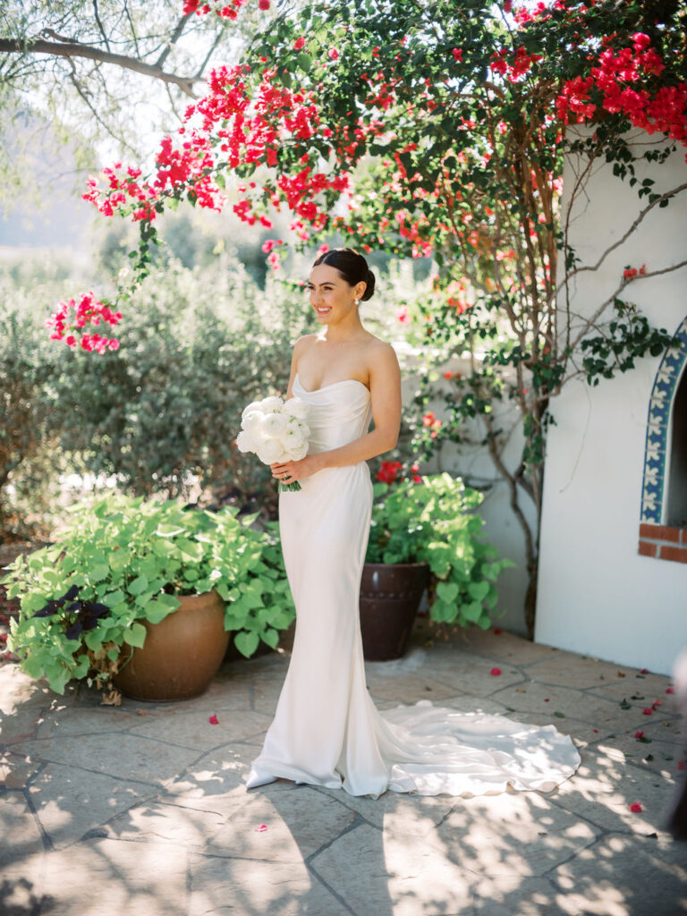 Bride standing on paved area under bougainvillea growing beside building, smiling and holding bouquet of white ranunculus flowers.