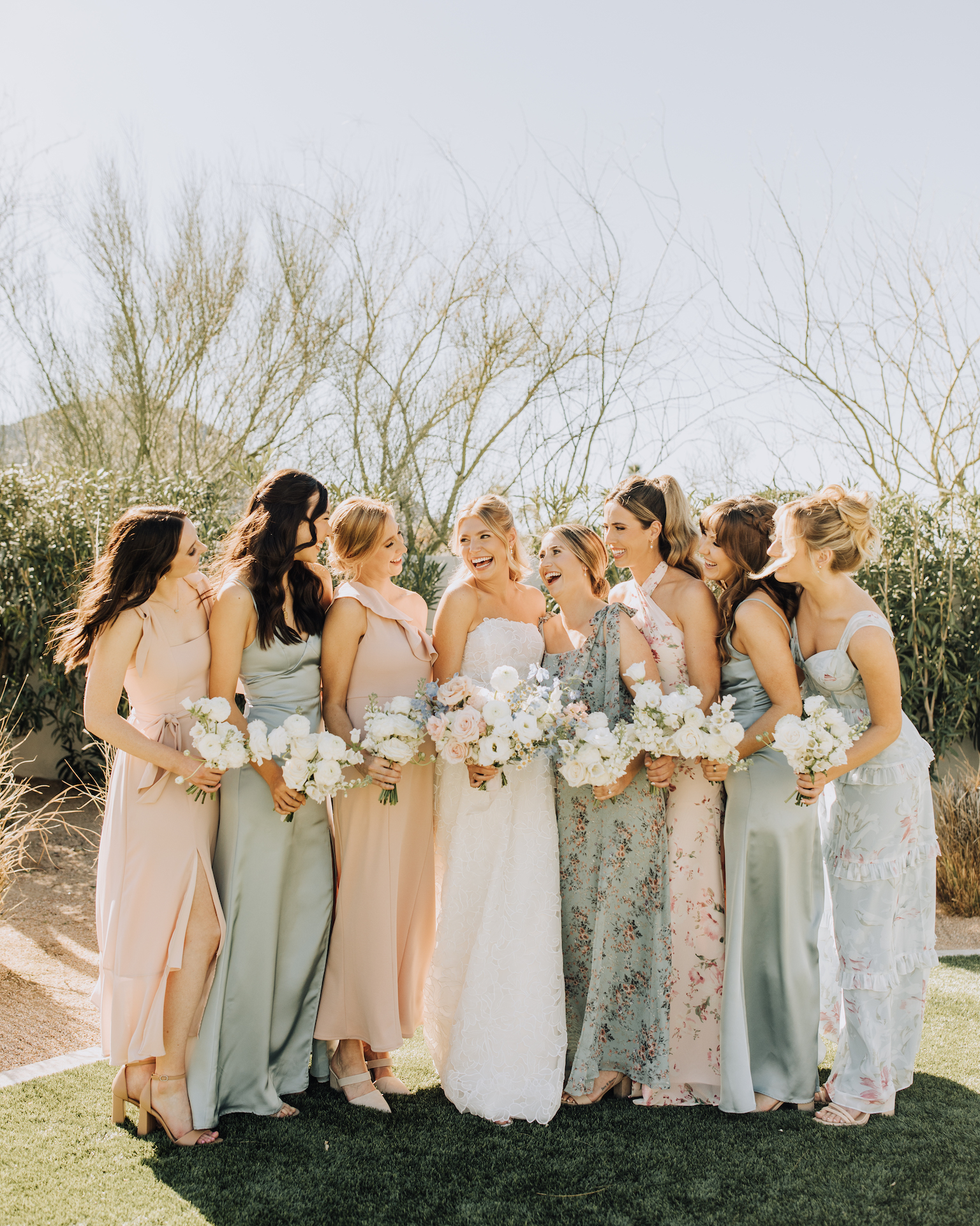 Bride with bridesmaids in varying shades of sage green and blush dresses all standing in a line with bouquets.