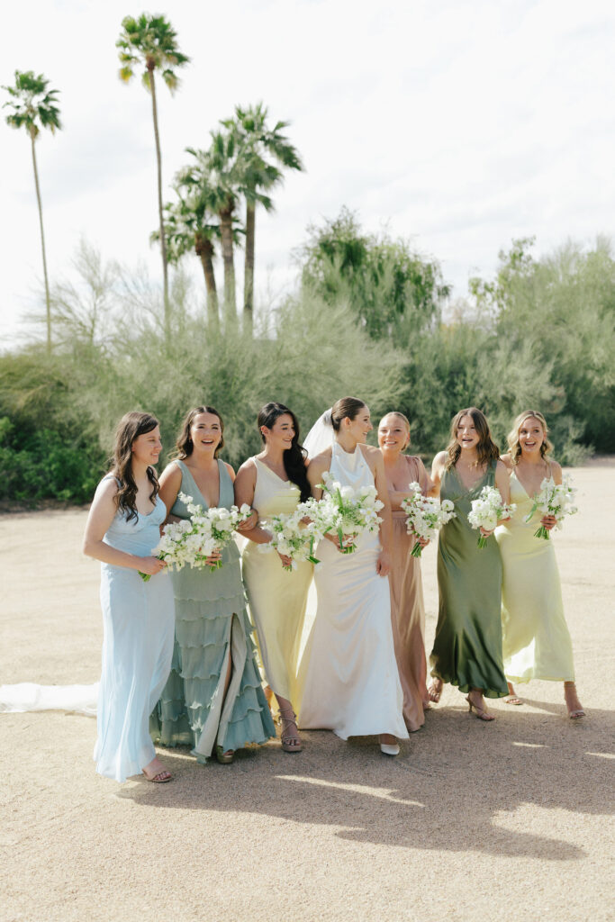 Bride walking in a row with bridesmaids wearing shades of yellow, blue, and green colors dresses in desert landscape.