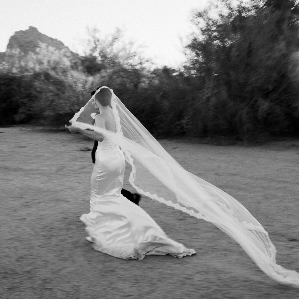 Bride and groom walking in desert landscape with bride's sheer long veil trailing behind.