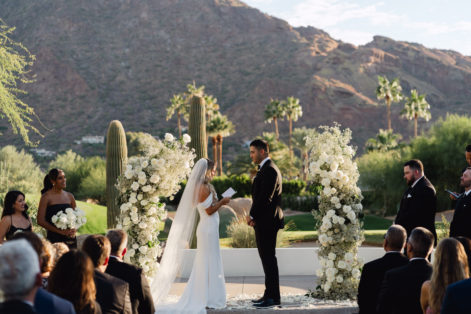 Bride reading vows to groom at outdoor wedding ceremony with white flowers framing them on either side.