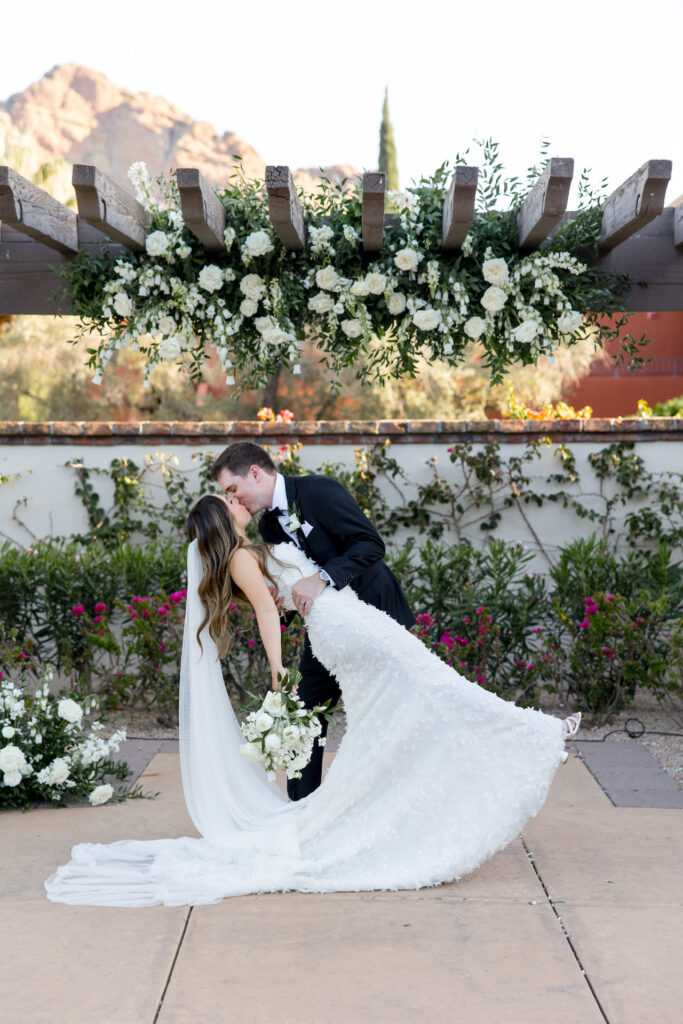 Groom dipping bride and kissing her under pergola with white floral and greenery installed on it.