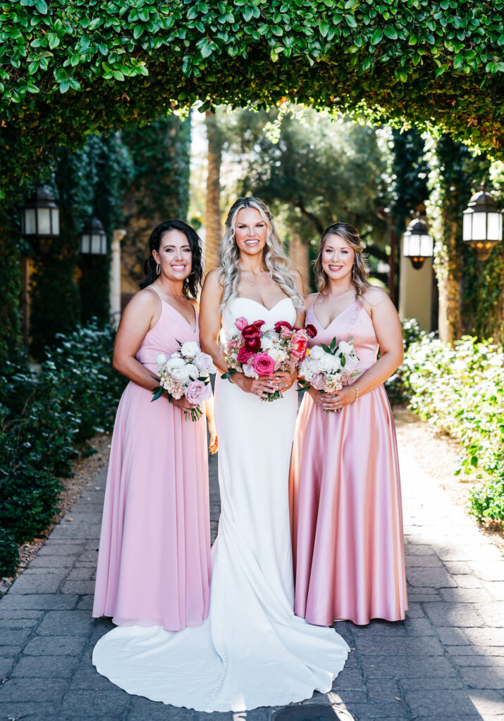 Bride in white standing between two women in pink dresses, all holding bouquets under an archway of greenery.