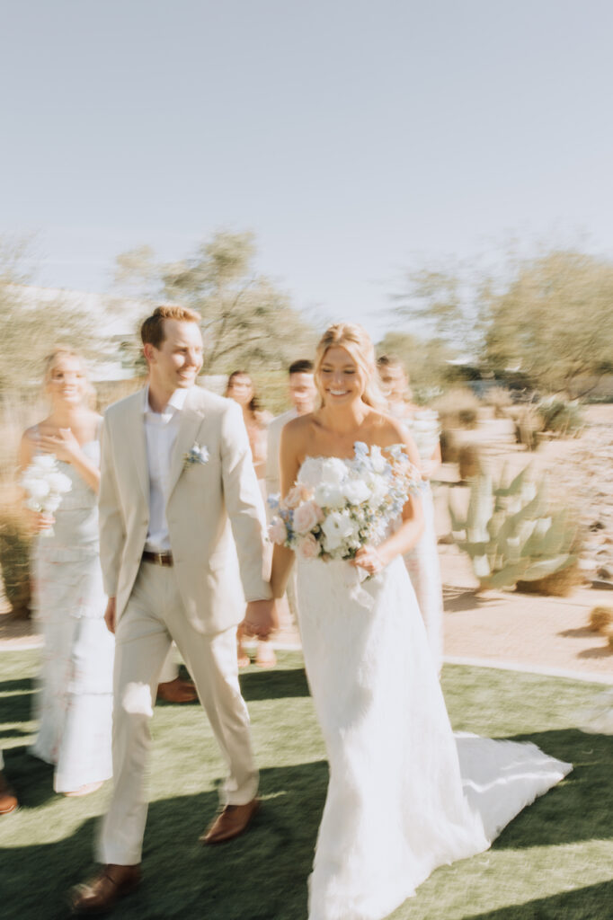 Blurry photo of bride and groom walking hand in hand with wedding party behind them and desert landscape in background.