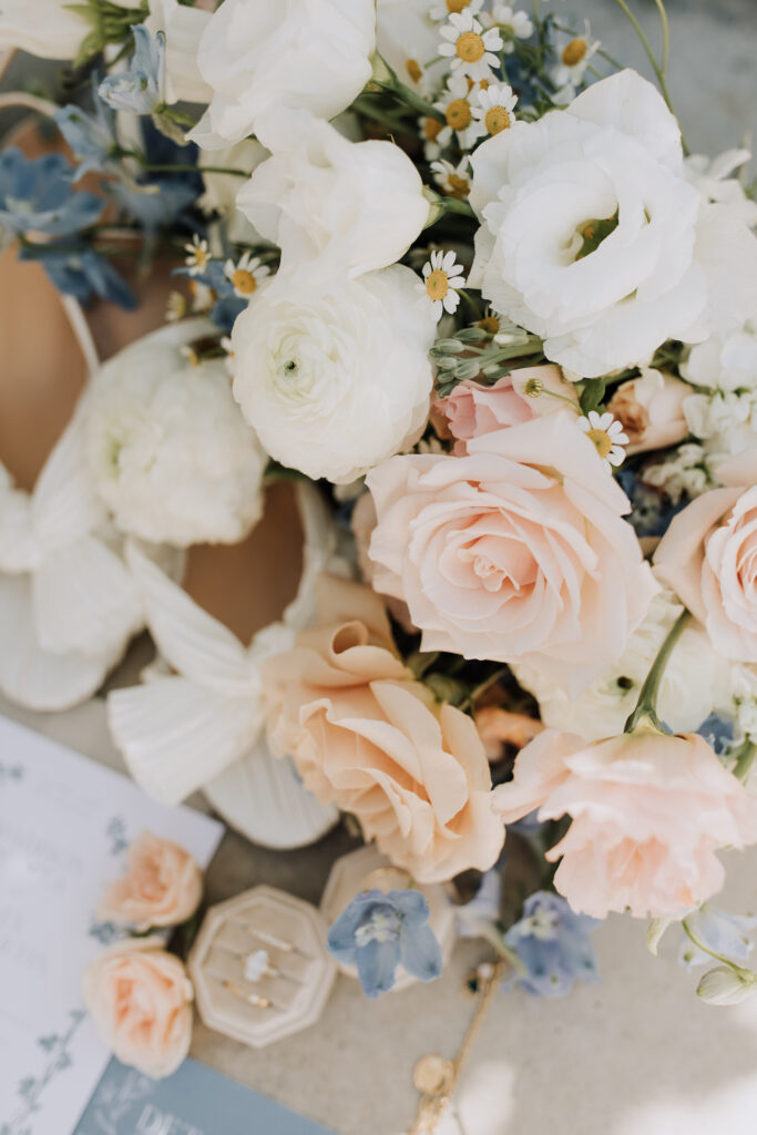 Beautiful wedding flowers bouquet of white, blush, and small amounts of blue flowers with bridal shoes and rings in background.