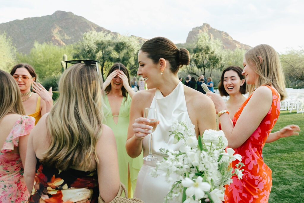 Bride standing around with other women dressed semi-formally at outdoor wedding ceremony, bride holding bouquet in one hand and champagne glass in other hand.