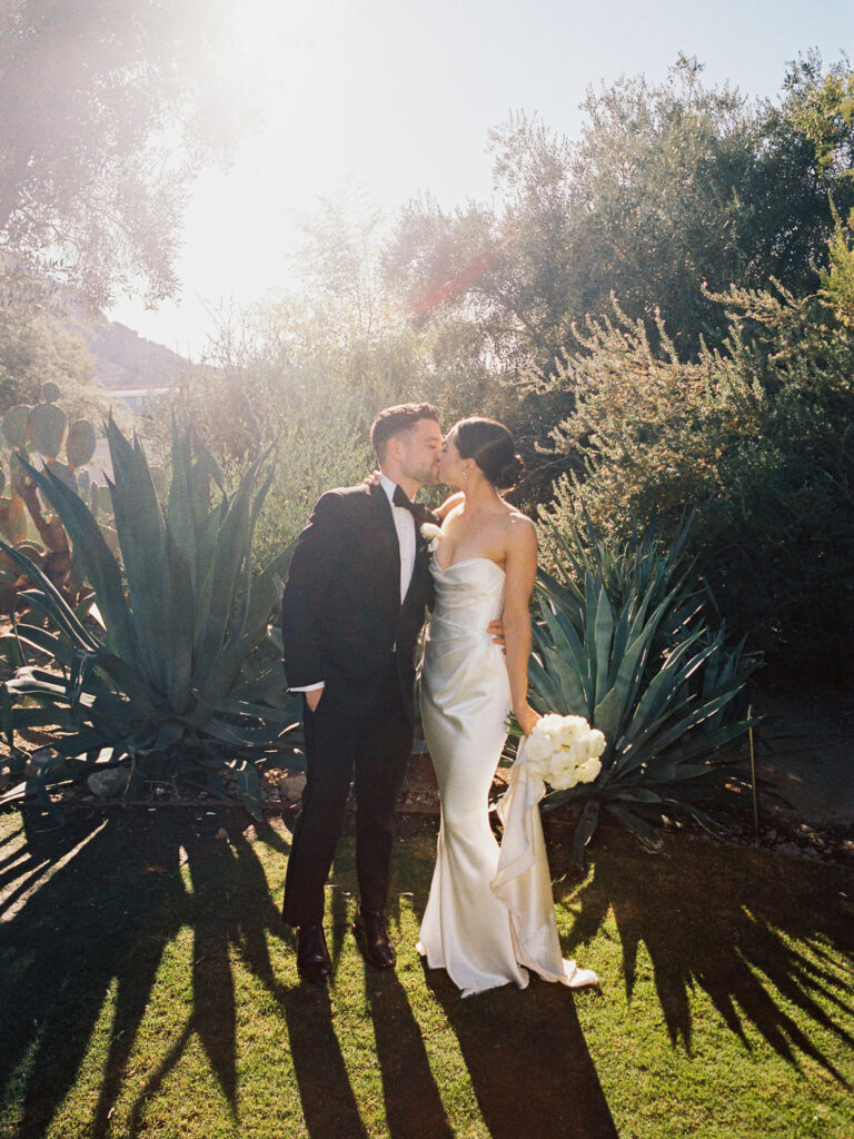 Bride and groom kissing, bride holding her white flowers bouquet down to side, standing in front of large agave looking plants.