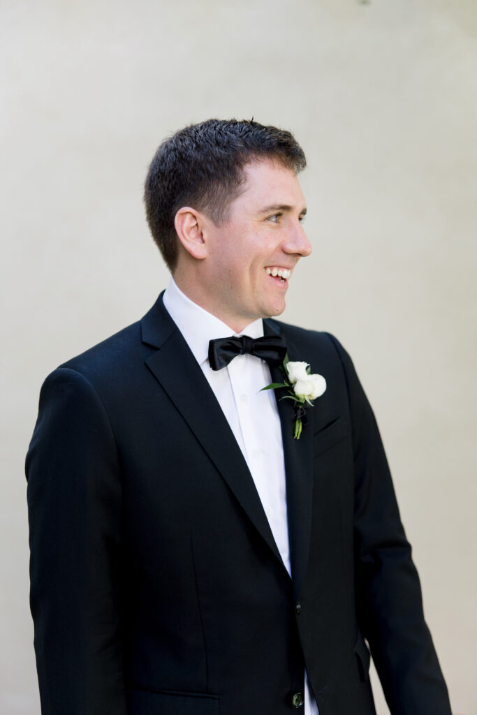 Groom in black suit smiling, looking to side.