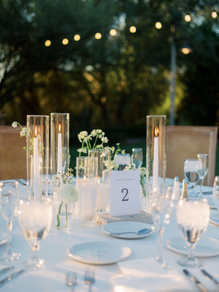 Wedding reception centerpiece design of taper candles with hurricanes over them and white flowers in bud vases.