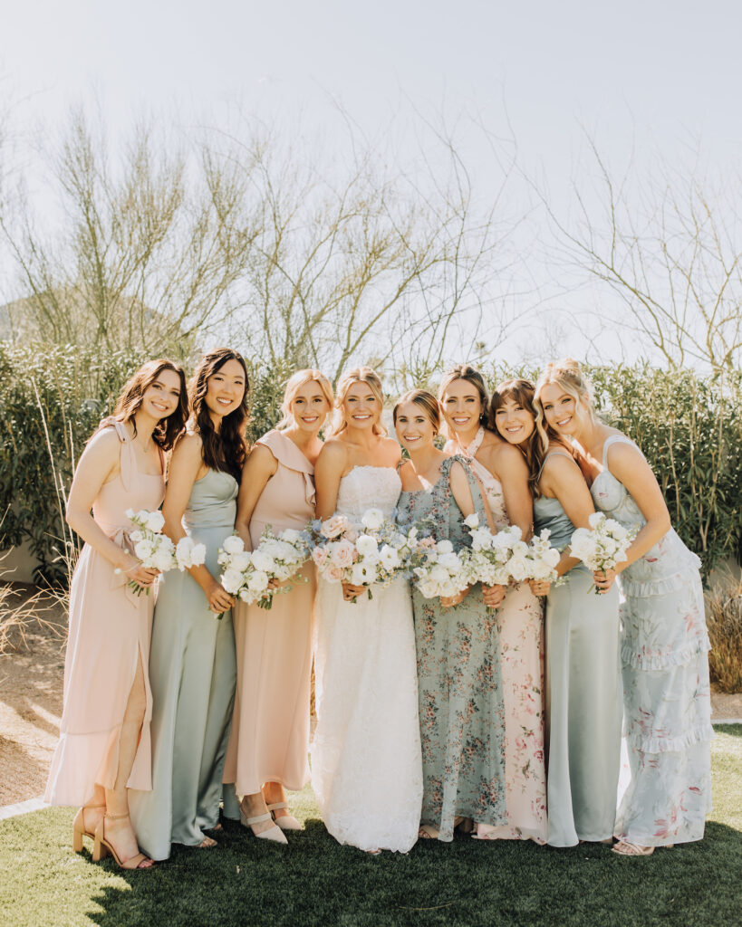 Bride with bridesmaids in varying shades of sage green and blush dresses all standing in a line with bouquets.