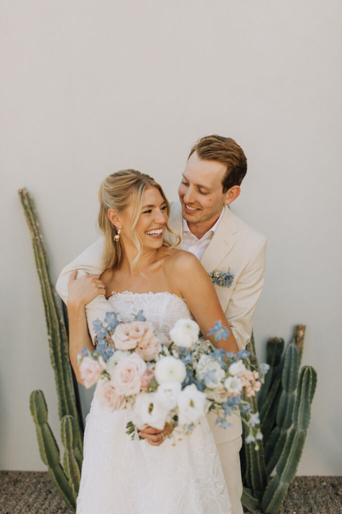 Broom standing behind bride with arms around her, both smiling, bride holding bouquet.