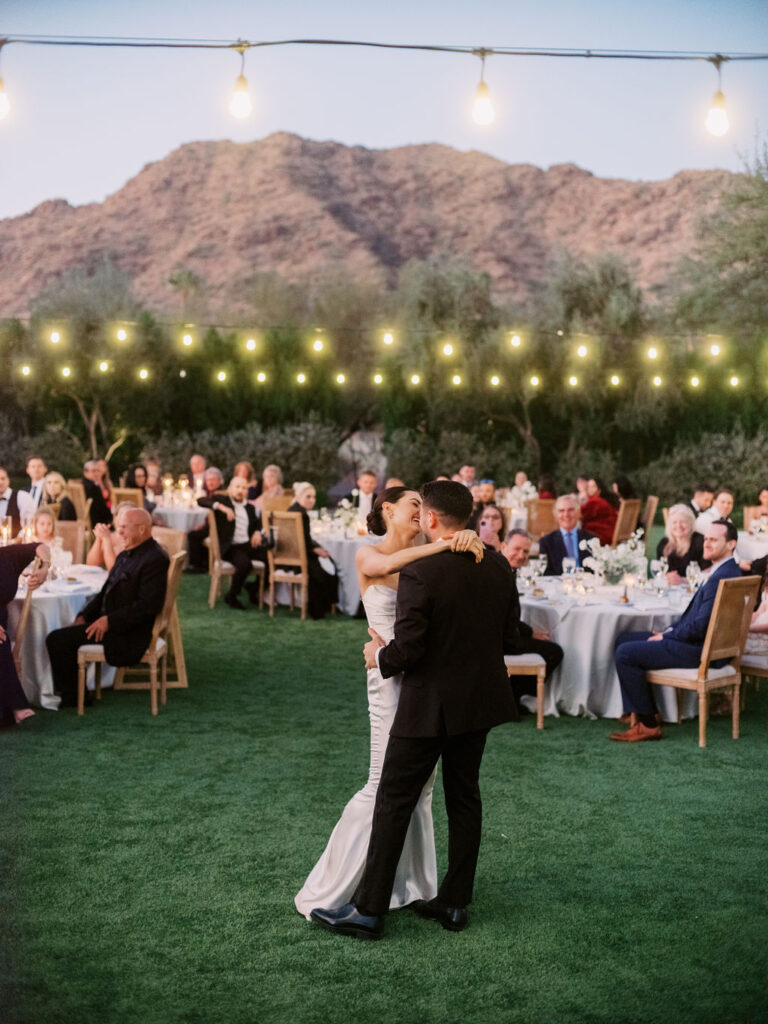 Bride and groom dancing on grass at outdoor reception with lights suspended overhead.