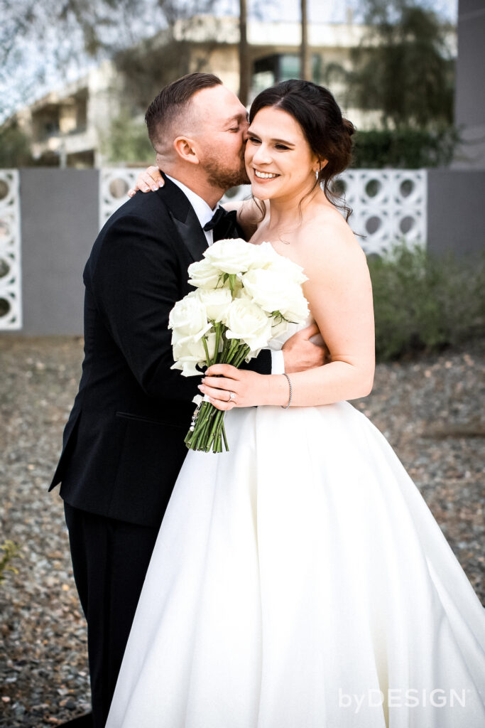 Groom kissing bride's cheek, embracing each other and smiling. Bride holding white roses bouquet.