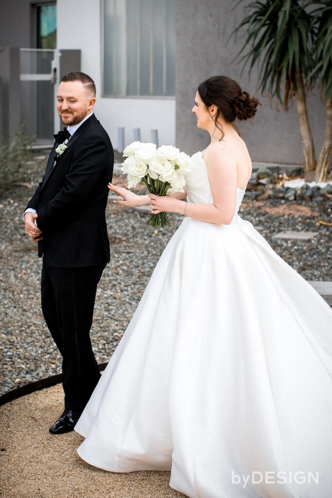 Wedding first look with groom turning around to see bride in white gown, holding white roses bouquet.