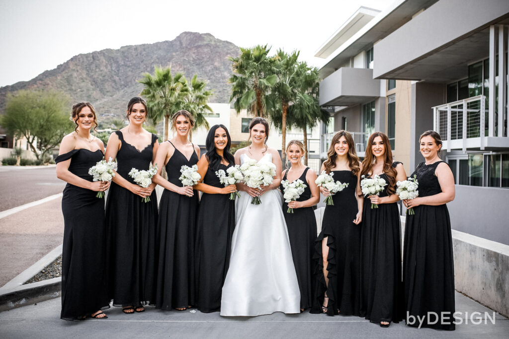 Bride standing in center of line of bridesmaids wearing different cuts of black dresses.