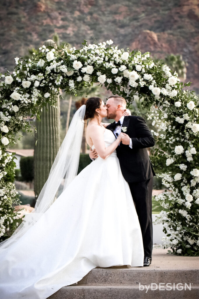 Bride and groom kissing under wedding ceremony arch of white flowers and greenery.