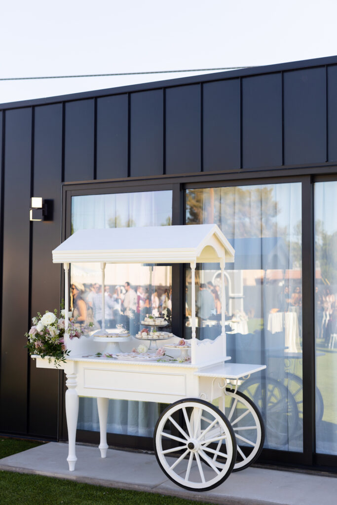 Dessert cart at baby shower with flowers placed on outside ledge of pink and white colors in wooden basket.