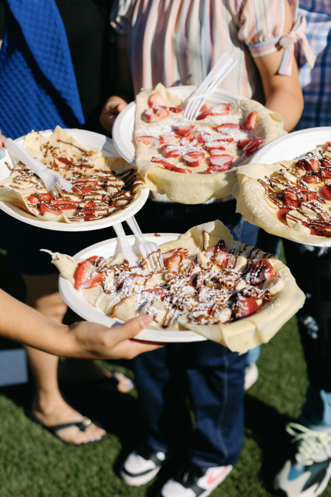 People holding crepes on paper plates with strawberries, chocolate sauce, and powdered sugar.