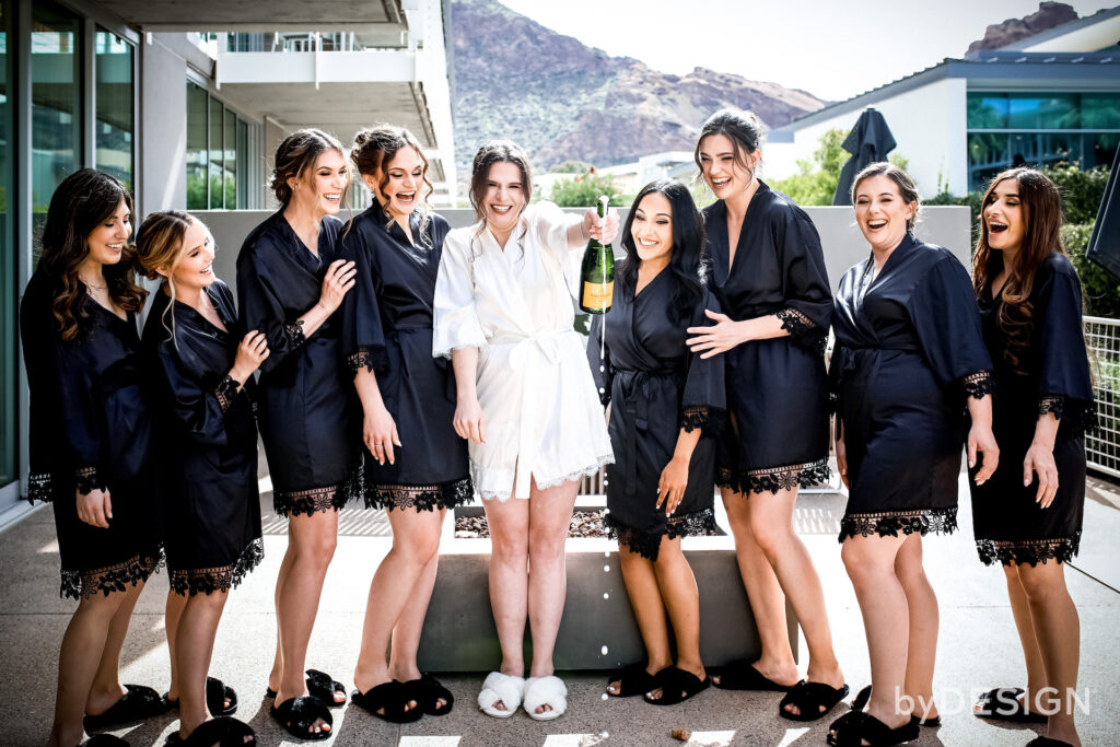 Bride opening champagne bottle on patio with bridesmaids standing on either side of her.