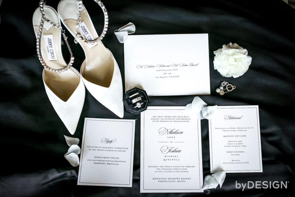 Wedding invitation flat lay, bridal shoes and jewelry on black backdrop.