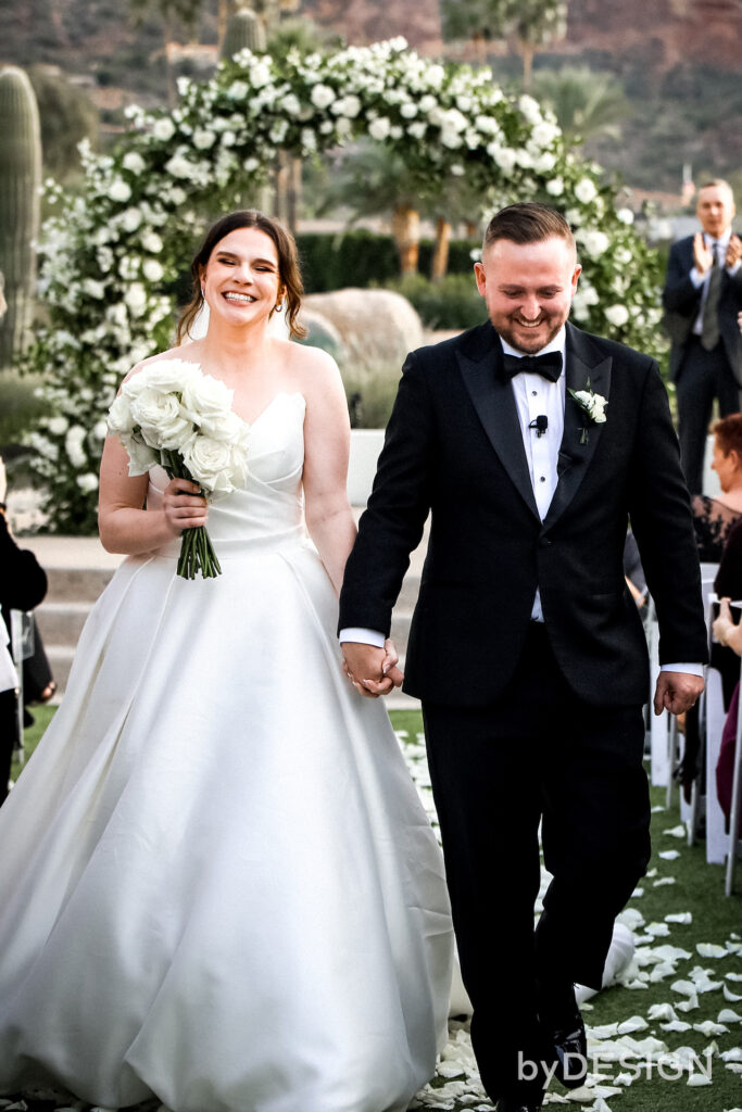 Bride and groom exiting down wedding ceremony aisle, smiling, holding hands.